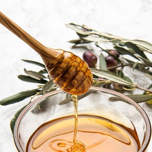 May include: A wooden honey dipper with a drizzling stream of honey into a clear glass bowl. Olive branches with green leaves and dark purple olives are in the background.