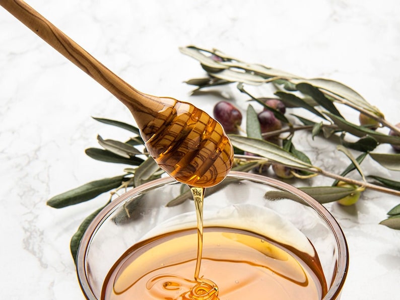 May include: A wooden honey dipper with a drizzling stream of honey into a clear glass bowl. Olive branches with green leaves and dark purple olives are in the background.