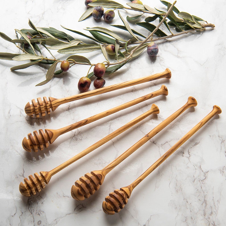May include: Six wooden honey dippers with a natural wood finish. The dippers have a long handle and a honeycomb-shaped scoop at the end. The dippers are arranged in a fan shape on a white marble surface.