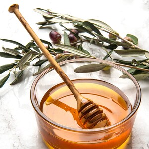 May include: A glass bowl filled with honey and a wooden honey dipper. Olive branches with green leaves and dark purple olives are in the background.