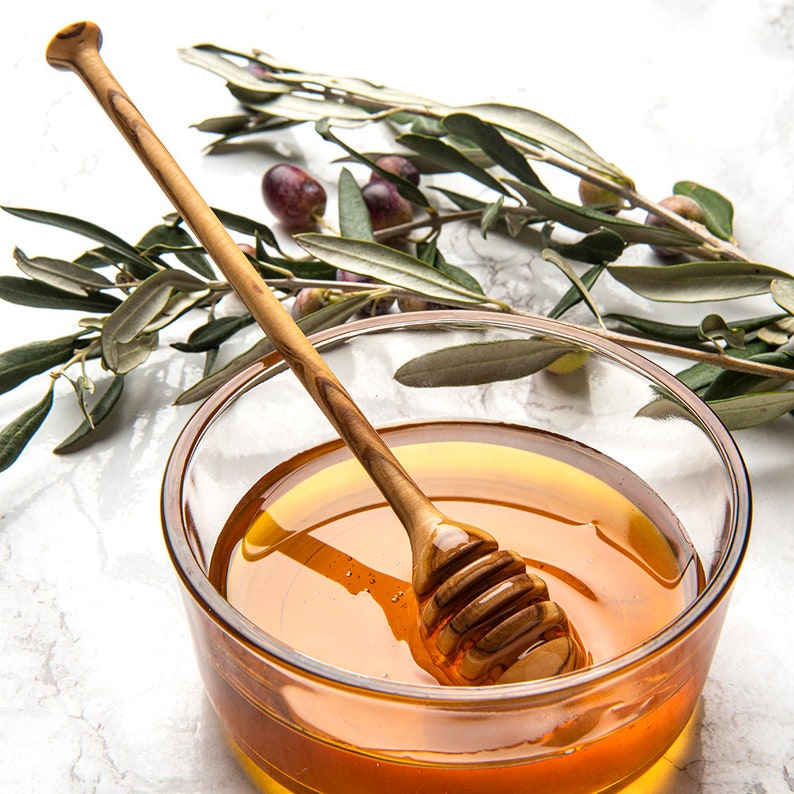May include: A glass bowl filled with honey and a wooden honey dipper. Olive branches with green leaves and dark purple olives are in the background.