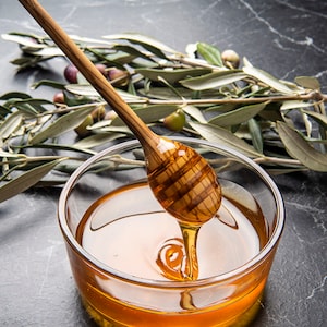 May include: A wooden honey dipper with a honeycomb design is dripping honey into a clear glass bowl. Olive branches with green leaves and small olives are in the background.
