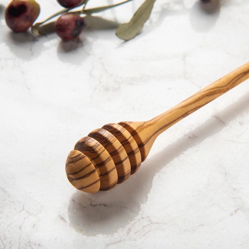 May include: A wooden honey dipper with a natural wood grain finish. The dipper has a long handle and a rounded, honeycomb-shaped head.