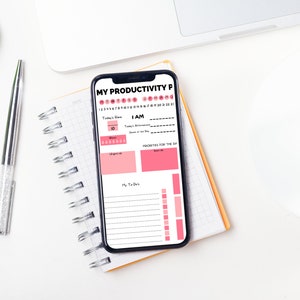 Op de afbeelding: Een witte smartphone met een roze en zwarte productiviteits-planner-app die openstaat op het scherm. De app bevat secties voor de focus van vandaag, Ik ben, prioriteiten voor de dag en mijn taken. De telefoon ligt op een geel en wit spiraalnotitieblok.