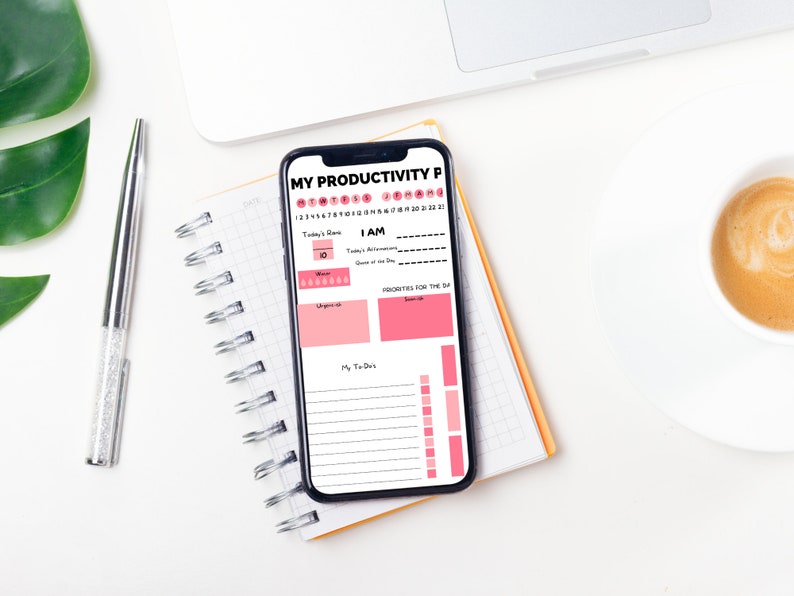 Op de afbeelding: Een witte smartphone met een roze en zwarte productiviteits-planner-app die openstaat op het scherm. De app bevat secties voor de focus van vandaag, Ik ben, prioriteiten voor de dag en mijn taken. De telefoon ligt op een geel en wit spiraalnotitieblok.