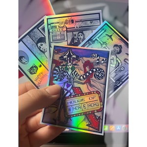 May include: A set of four iridescent stickers featuring illustrations of horror movie characters. The stickers are printed on a holographic background and feature a black and white striped design. The text on one sticker reads "House of Horrors".