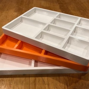 May include: Three stacked plastic trays with compartments. The top tray is white and has nine compartments. The middle tray is orange and has four compartments. The bottom tray is white and has four compartments.