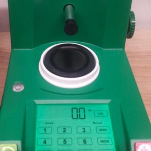 May include: A green digital scale with a black and white weighing platform. The scale has a digital display showing "00" and buttons labeled "1", "2", "3", "4", "5", "6", "Zero", "Mode", and "Cal".