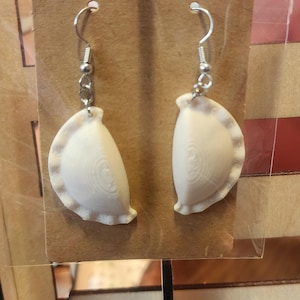 May include: A pair of white, handmade earrings shaped like empanadas. The earrings are hanging from silver hooks.