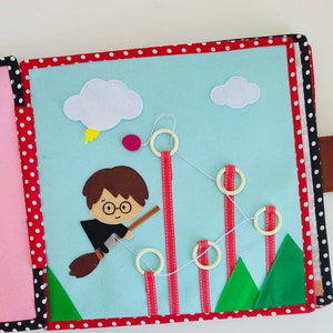 May include: A felt book for children featuring a character flying on a broomstick with a red and white striped ribbon and a string of rings. The book has a blue background with clouds and green trees.