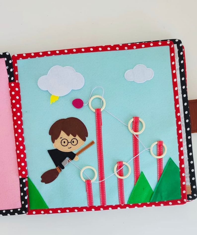 May include: A felt book for children featuring a character flying on a broomstick with a red and white striped ribbon and a string of rings. The book has a blue background with clouds and green trees.