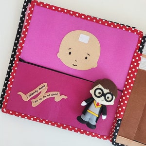 May include: A pink felt book with red polka dots on the cover. The book has a felt character of a baby with a white bandage on its forehead. The book also has a felt character of a boy with glasses and a brown robe. The book has a felt banner with the text "I solemnly swear I am up to no good".