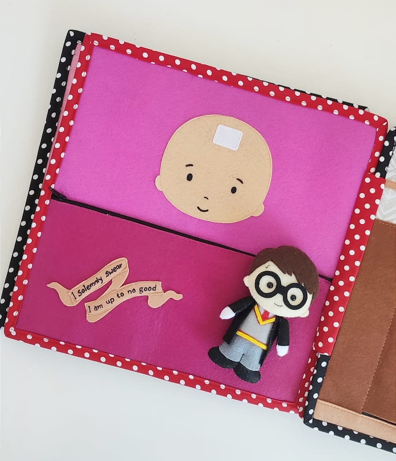 May include: A pink felt book with red polka dots on the cover. The book has a felt character of a baby with a white bandage on its forehead. The book also has a felt character of a boy with glasses and a brown robe. The book has a felt banner with the text "I solemnly swear I am up to no good".