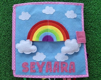 Può includere: Un libro di feltro blu con un arcobaleno, nuvole e il nome "SEYAAARA" in lettere di feltro rosa. Il libro ha pois rosa intorno ai bordi e una patta di feltro rosa sul lato.