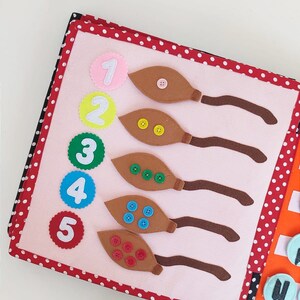 May include: A pink felt book with red polka dots on the cover. The book has five pages with numbers 1 through 5 and brown felt leaves with buttons on each page.
