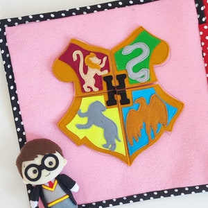 May include: A pink felt book with a felt Harry Potter house crest on the cover. The crest is made up of four different colored squares, each with a different animal. The book is surrounded by a black and white polka dot border.