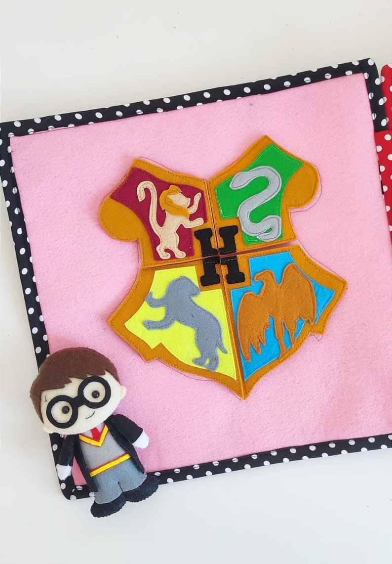 May include: A pink felt book with a felt Harry Potter house crest on the cover. The crest is made up of four different colored squares, each with a different animal. The book is surrounded by a black and white polka dot border.