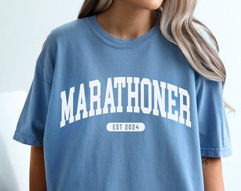 May include: A light blue t-shirt with the word "MARATHONER" in white block letters. Below the word is "EST 2024". The shirt is a washed denim style.