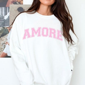 May include: White sweatshirt with the word "AMORE" printed in pink on the front.