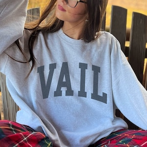 May include: A person wearing a light gray sweatshirt with the word "VAIL" printed in black on the front. They are sitting on a wooden bench wearing red and black plaid pants.