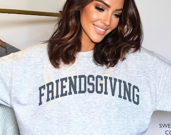 May include: A light gray sweatshirt with the word "FRIENDSGIVING" in dark gray block letters. The sweatshirt is paired with white sweatpants. The text "SWEATSHIRT COLOR ASH" is in the lower right corner.