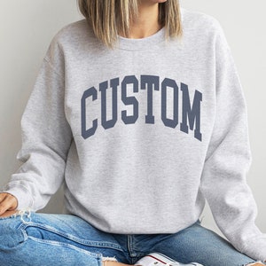 May include: A gray sweatshirt with the word "CUSTOM" printed in a navy blue arch across the front.