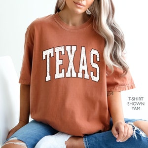 May include: A burnt orange t-shirt with a white distressed "TEXAS" graphic. The t-shirt is shown on a model wearing blue jeans.