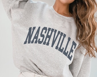 May include: Ash grey sweatshirt with the word "NASHVILLE" arched across the chest in navy blue lettering. The sweatshirt is a crew neck style. The text "SWEATSHIRT COLOR ASH GREY" is in the lower left corner.