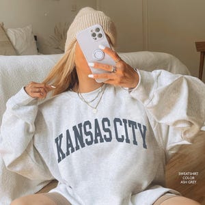Puede incluir: Una sudadera gris claro con el texto "KANSAS CITY" en letras negras. La sudadera la lleva puesta una persona sentada en un suelo de madera. La sudadera es de corte holgado y tiene cuello redondo.