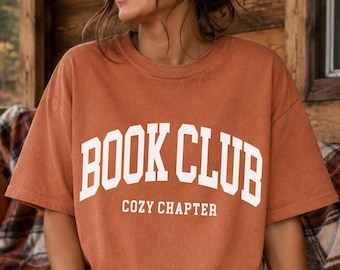 May include: A rust-colored short-sleeve t-shirt featuring the white text "BOOK CLUB" in a large arched collegiate font, with the smaller white text "COZY CHAPTER" printed directly underneath. The shirt has a crew neckline and a relaxed fit.
