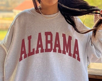 May include: A gray sweatshirt with the word "ALABAMA" in red letters across the front. The model is wearing the sweatshirt and has long brown hair. The text "MODEL SIZED UP 2 SIZES FOR THIS OVERSIZED FIT!" is in a red circle on the bottom right of the image.