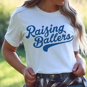 May include: White t-shirt with blue text that reads "Raising Ballers" in a retro font style. The text has a baseball-style arc underneath it.
