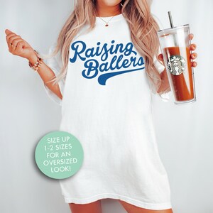 May include: White oversized t-shirt with blue text that reads "Raising Ballers". The shirt is being worn by a person holding a clear tumbler with a brown beverage. A light blue circle with white text says "Size up 1-2 sizes for an oversized look!"
