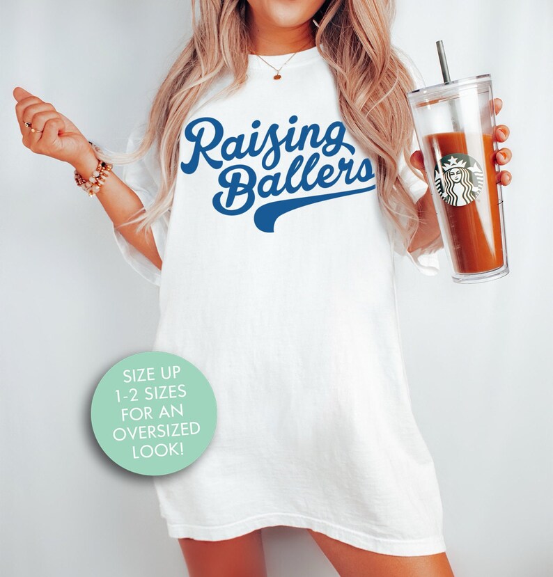 May include: White oversized t-shirt with blue text that reads "Raising Ballers". The shirt is being worn by a person holding a clear tumbler with a brown beverage. A light blue circle with white text says "Size up 1-2 sizes for an oversized look!"