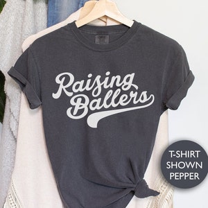 May include: A dark gray t-shirt with the text "Raising Ballers" in white lettering. The text is in a retro baseball font with a white baseball-style arc underneath.