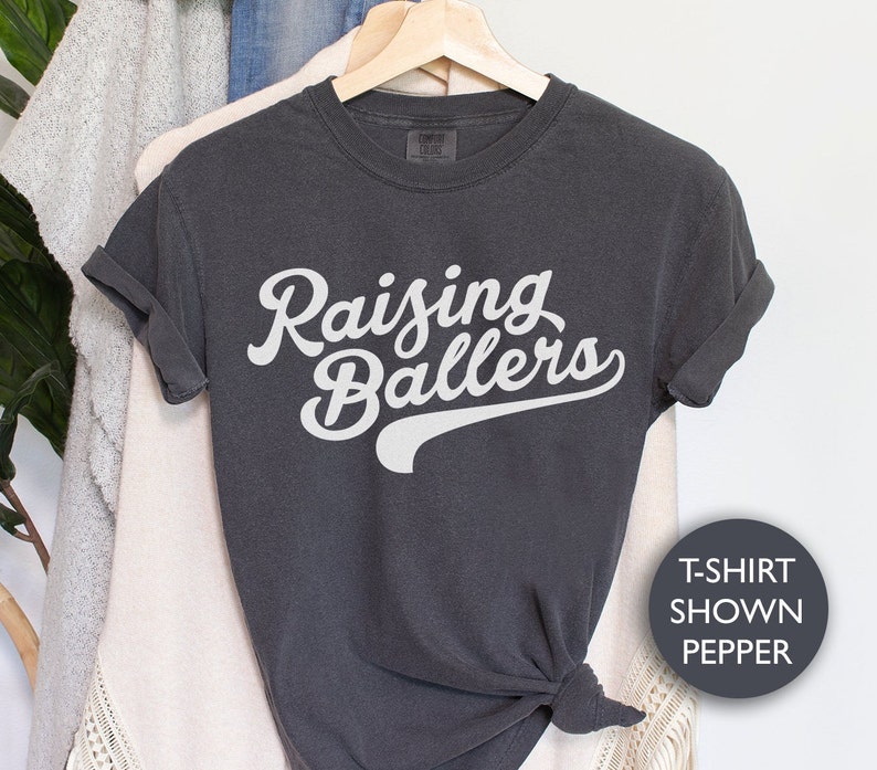 May include: A dark gray t-shirt with the text "Raising Ballers" in white lettering. The text is in a retro baseball font with a white baseball-style arc underneath.
