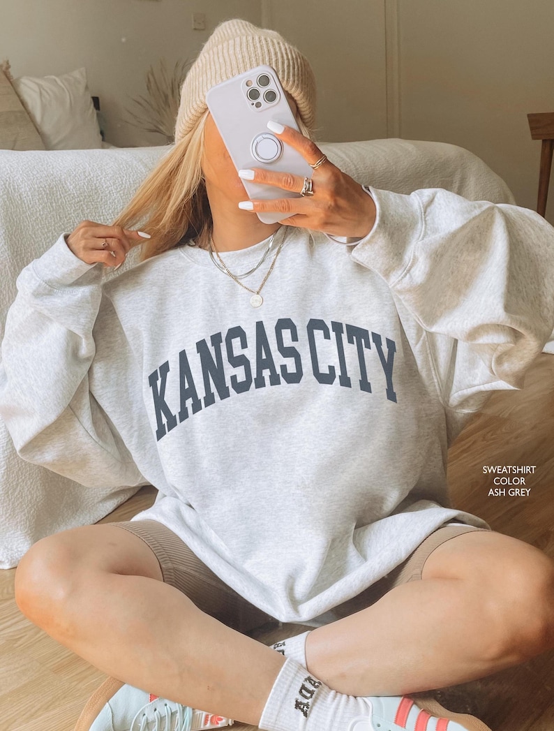 Puede incluir: Una sudadera gris claro con el texto "KANSAS CITY" en letras negras. La sudadera la lleva puesta una persona sentada en un suelo de madera. La sudadera es de corte holgado y tiene cuello redondo.