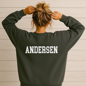 Może przedstawiać: Ciemnozielona bluza z okrągłym dekoltem z napisem "ANDERSEN" pogrubionymi, białymi literami na plecach. Bluza jest w jednolitym kolorze i ma długie rękawy. Osoba na zdjęciu ma na sobie bluzę.