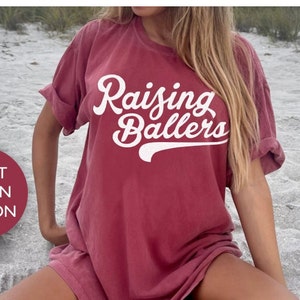 May include: A crimson t-shirt with the text "Raising Ballers" in white cursive lettering with a baseball stitch design.