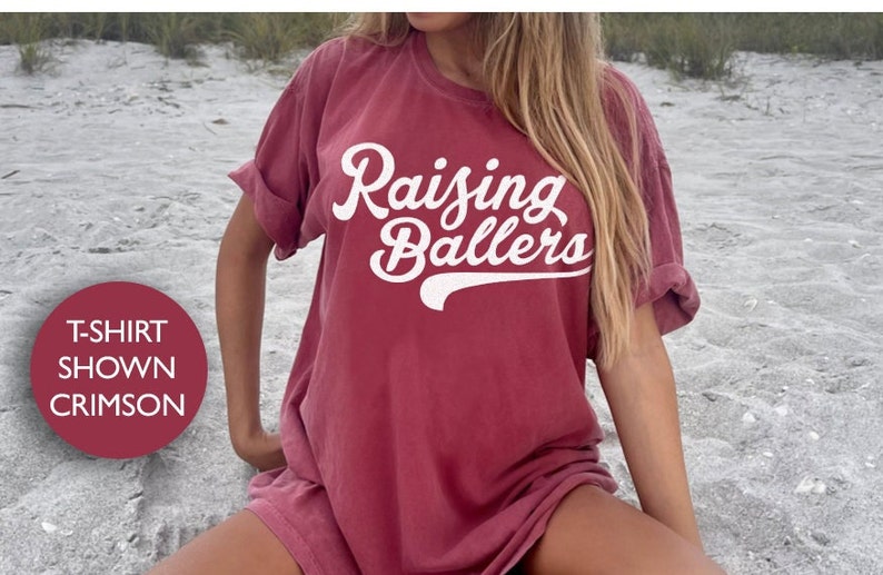May include: A crimson t-shirt with the text "Raising Ballers" in white cursive lettering with a baseball stitch design.