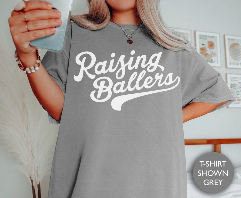 May include: A gray t-shirt with the text "Raising Ballers" in white. The shirt is shown on a model.