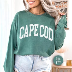 May include: A light green sweatshirt with the text "CAPE COD" in white letters. The sweatshirt is made of a soft, comfortable material and has a relaxed fit.
