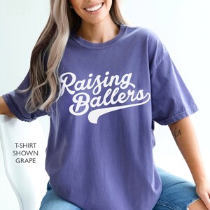 May include: A purple t-shirt with the text "Raising Ballers" in white cursive lettering with a white baseball-style arc underneath the text.