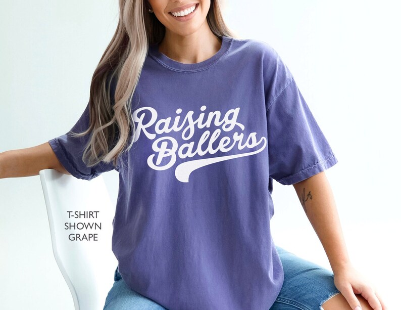 May include: A purple t-shirt with the text "Raising Ballers" in white cursive lettering with a white baseball-style arc underneath the text.