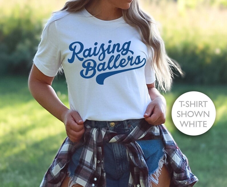 May include: White t-shirt with blue text that reads "Raising Ballers" in a retro font style. The text has a baseball-style arc underneath it.