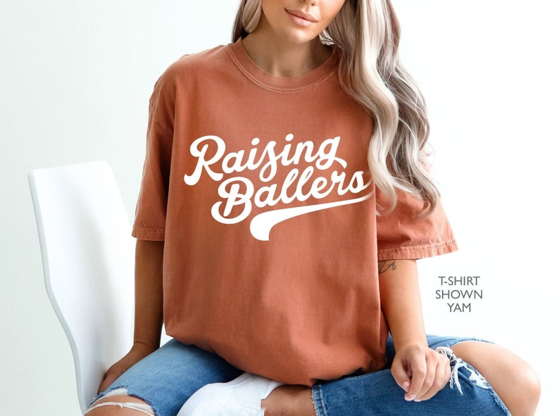 May include: A rust-colored t-shirt with the text "Raising Ballers" in white script with a white baseball-style arc underneath.