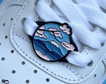 May include: A white trainer with a black and blue shoelace charm featuring a mountain landscape with pink clouds.