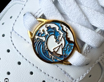 May include: A gold-coloured shoe charm with a blue and white wave design and a sailboat. The charm is on white shoelaces and a white leather shoe. The brand name "DOOSTOMZ" is visible in the lower left corner.