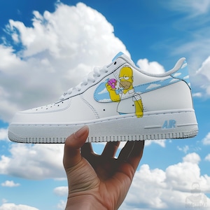 Puede incluir: Zapatillas Air Force 1 blancas con una imagen de dibujos animados de Homer Simpson sosteniendo una rosquilla. La zapatilla tiene un logotipo azul claro con detalles de nubes y la palabra "AIR" en azul. El fondo es un cielo azul nublado.