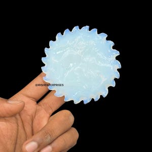 Puede incluir: Una gema opalescente, redonda y translúcida con borde festoneado, sobre fondo negro. La piedra tiene un sutil patrón interno y un contorno azul claro. El texto "IDRISHENTERPRISES" es visible.